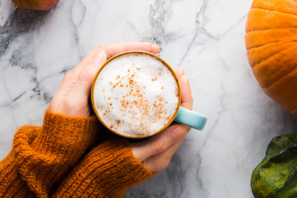 Autumn moody background with mug of latte coffee and pumpkins on marble table. Flat lay in fall colors. Female hands in cozy sweater